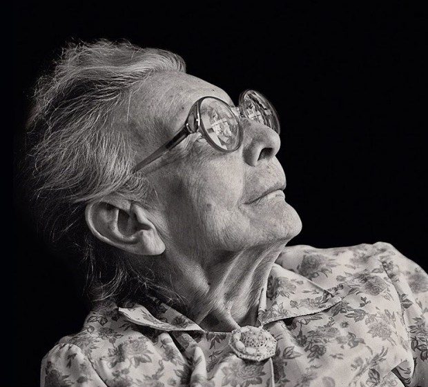 A imagem é uma foto de Nise da Silveira. Ela está olhando para cima e para direita. Seus cabelos grisalhos estão presos e ela está usando óculos de lentes redondas e um vestido florido.