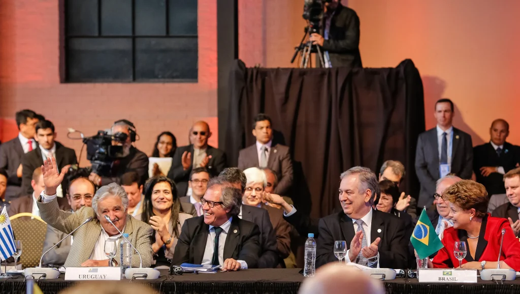 No primeiro plano estão José Mujica e Dilma Rousseff, ex-presidentes do Uruguai e do Brasil, respectivamente. Atrás deles estão diversas pessoas também sentadas.