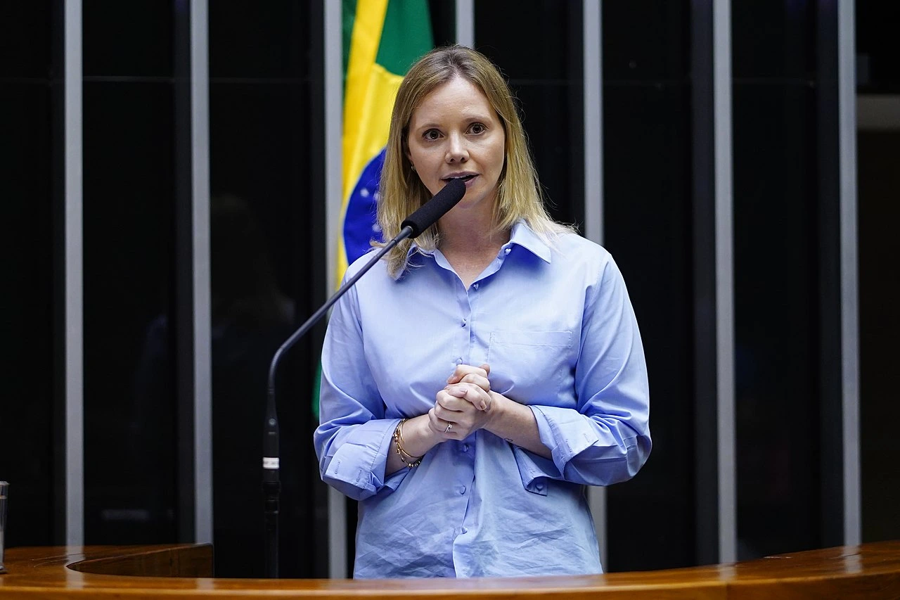 Dep. Dani Cunha, autora do projeto para criminalizar a discriminação contra pessoas politicamente expostas Deputada Dani Cunha (UNIAO - RJ) no plenário da Câmara dos Deputados, onde apresentou o PL 2720/2023 para criminalizar a discriminação contra pessoas politicamente expostas. Usa camisa social azul, em fundo preto com bandeira do Brasil.