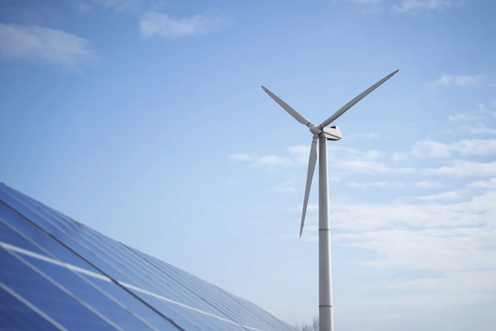 Foto com fundo do céu, à esquerda uma placa de produção de energia solar e à direita produção de energia eólica, evidenciando a diversidade de produção da energia limpa
