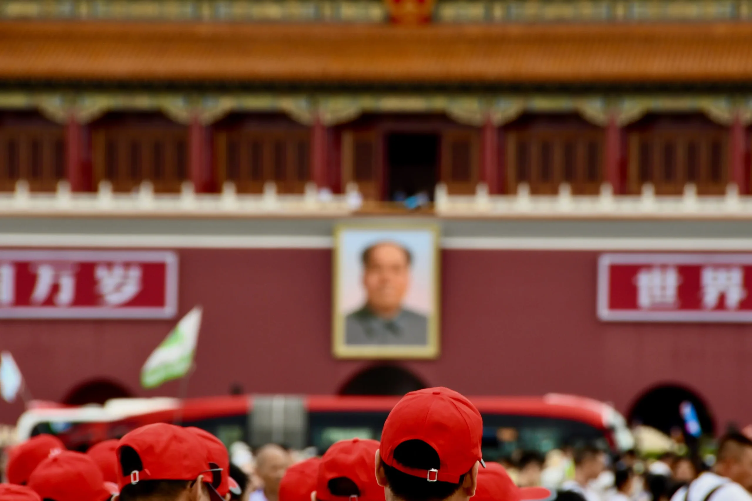 a imagem apresenta, em um plano principal, um prédio com uma imagem de Mao Tsé-Tung. Abaixo, percebe-se uma multidão em deslocamento. No primeiro plano da imagem, as pessoas usam bonés vermelhos.