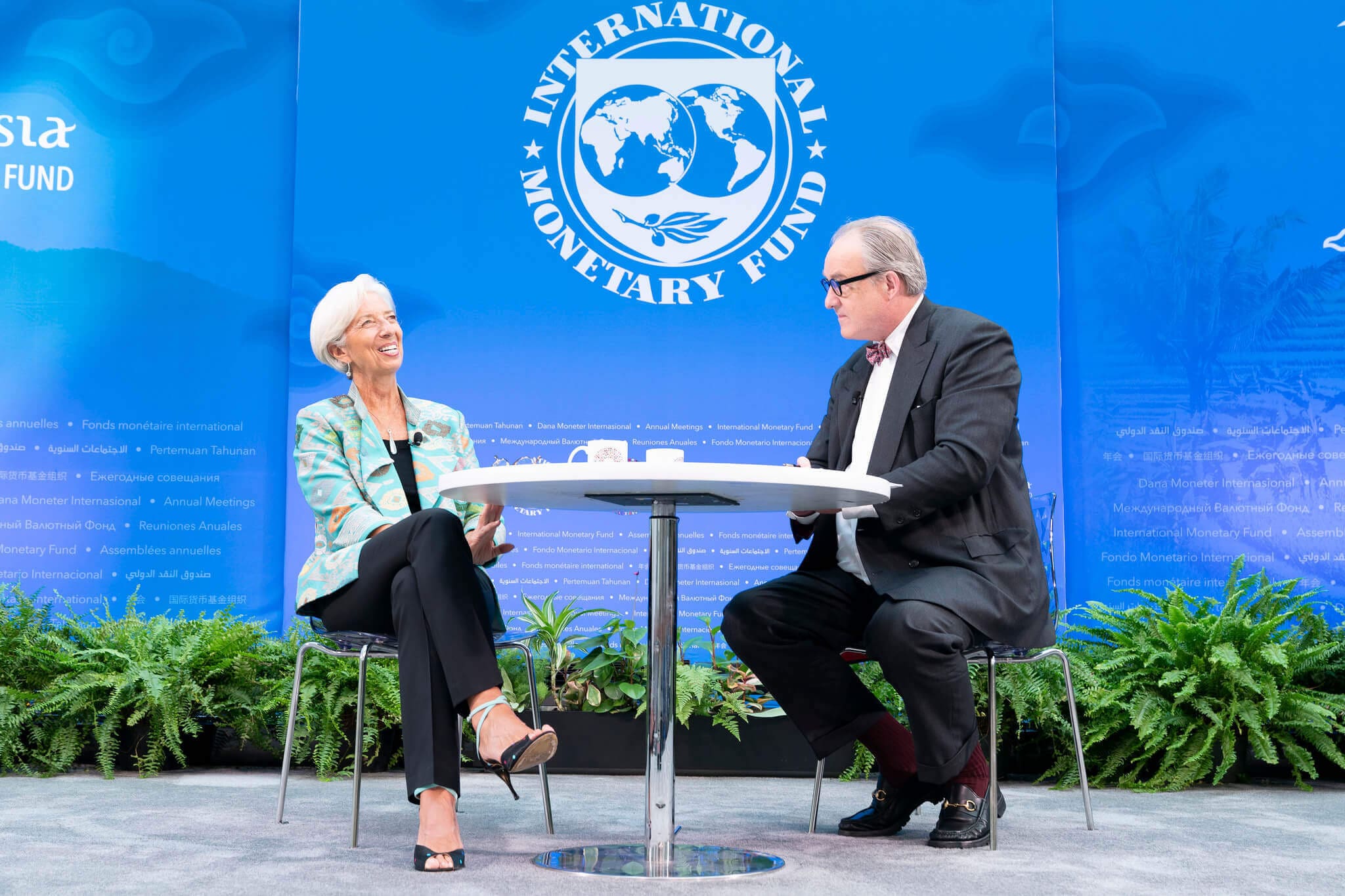 Dois diretores do Fundo Monetário Internacional sentados em uma mesa conversando. Ao fundo o logo do FMI.