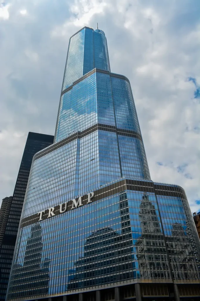 Prédio alto espelhado com o nome Trump escrito na entrada contra um céu de nuvens. Texto sobre Donald Trump.