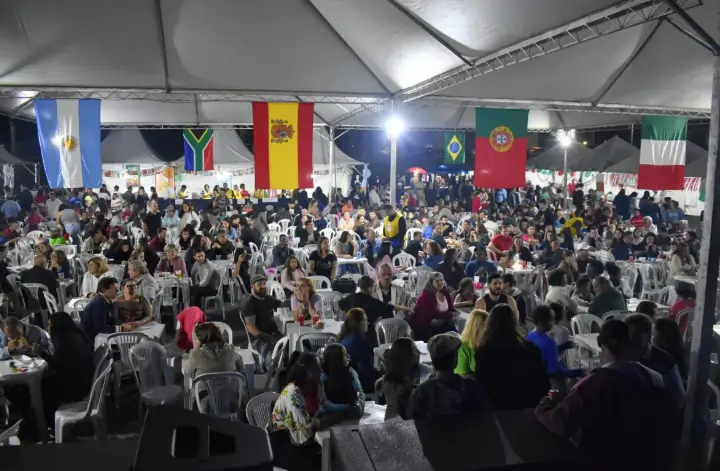 Imagem de várias pessoas reunidas em um salão, sentadas em volta de cadeiras de plástico, em cadeiras de plástico, com várias bandeiras de nações diferentes penduradas pelo salão. Texto: Você sabe o que é multiculturalismo? 