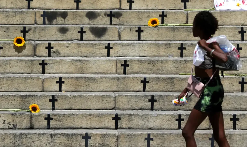 Foto de uma mulher negra passando em frente a uma escadaria com várias cruzes e girassois. Texto: Lei do Feminicídio — 10 anos de avanços e desafios
