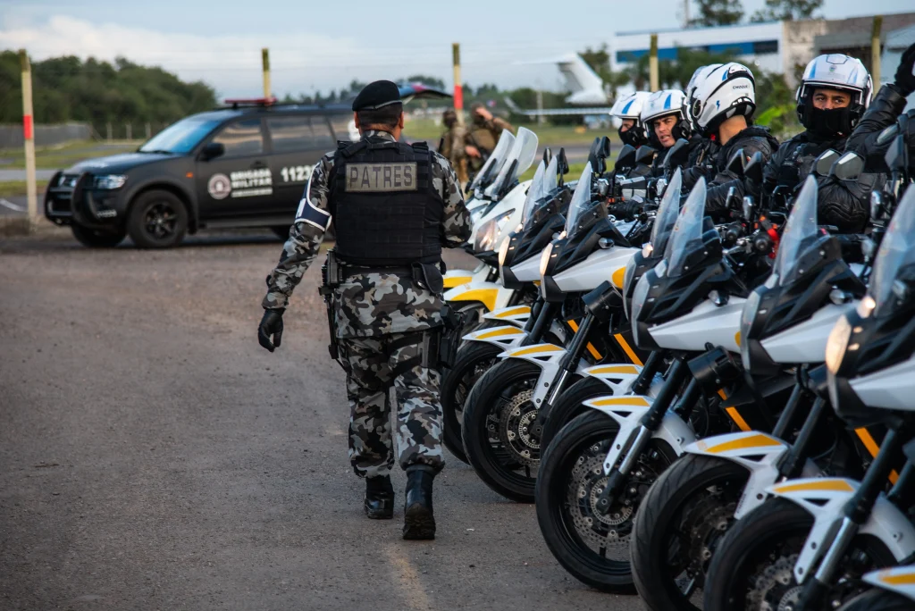 Policial Militar de costas caminha em direção a equipe de motociclistas, alinhados ao lado de suas motos em pátio externo.