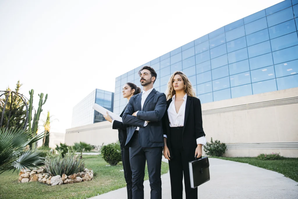 Imagem de três pessoas jovens duas mulheres brancas e um homem, usando roupas sociais em frente um prédio empresarial espelhado