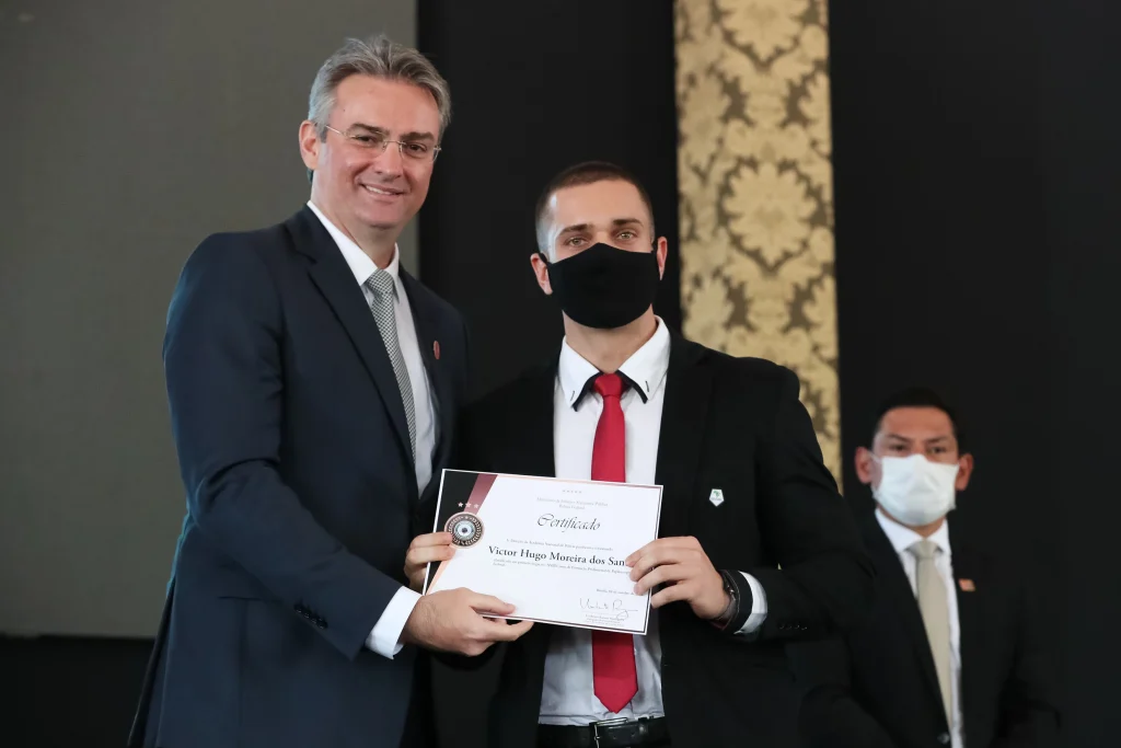 Homem branco de meia-idade, de terno e óculos, entrega um certificado a um formando de beca e máscara pretas; ambos posam e sorriem para a foto.