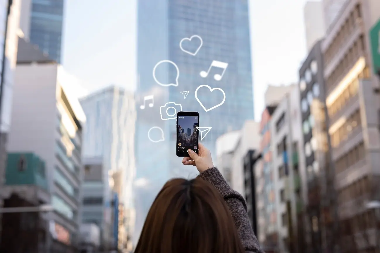 Pessoa vista de costas segura um smartphone erguido, fotografando ou gravando prédios altos em uma área urbana. Ícones gráficos de redes sociais, como coração, câmera, balão de conversa e notas musicais, aparecem sobrepostos à imagem, sugerindo produção ou compartilhamento de conteúdo digital.
